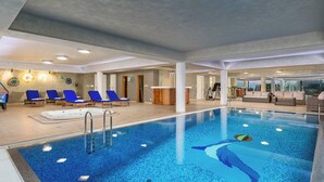 A large indoor swimming pool with blue mosaic tiles, surrounded by lounge chairs and a jacuzzi.