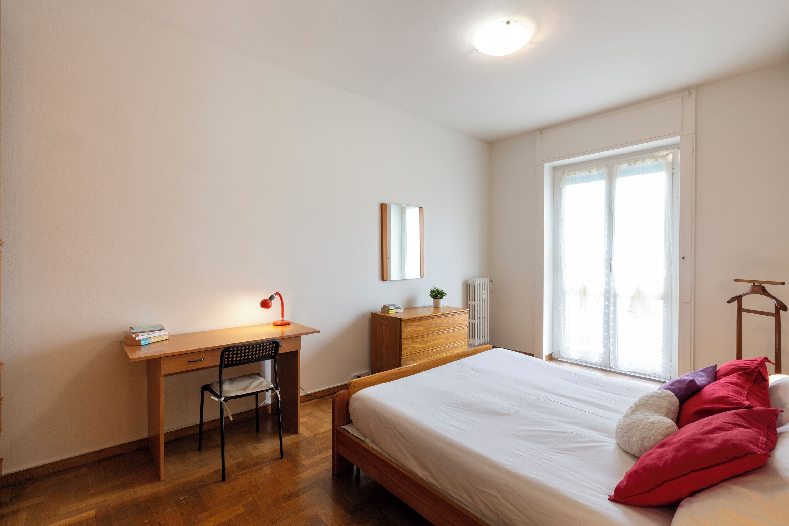 A bedroom with a wooden desk, chair, and a bed with white bedding and red pillows.