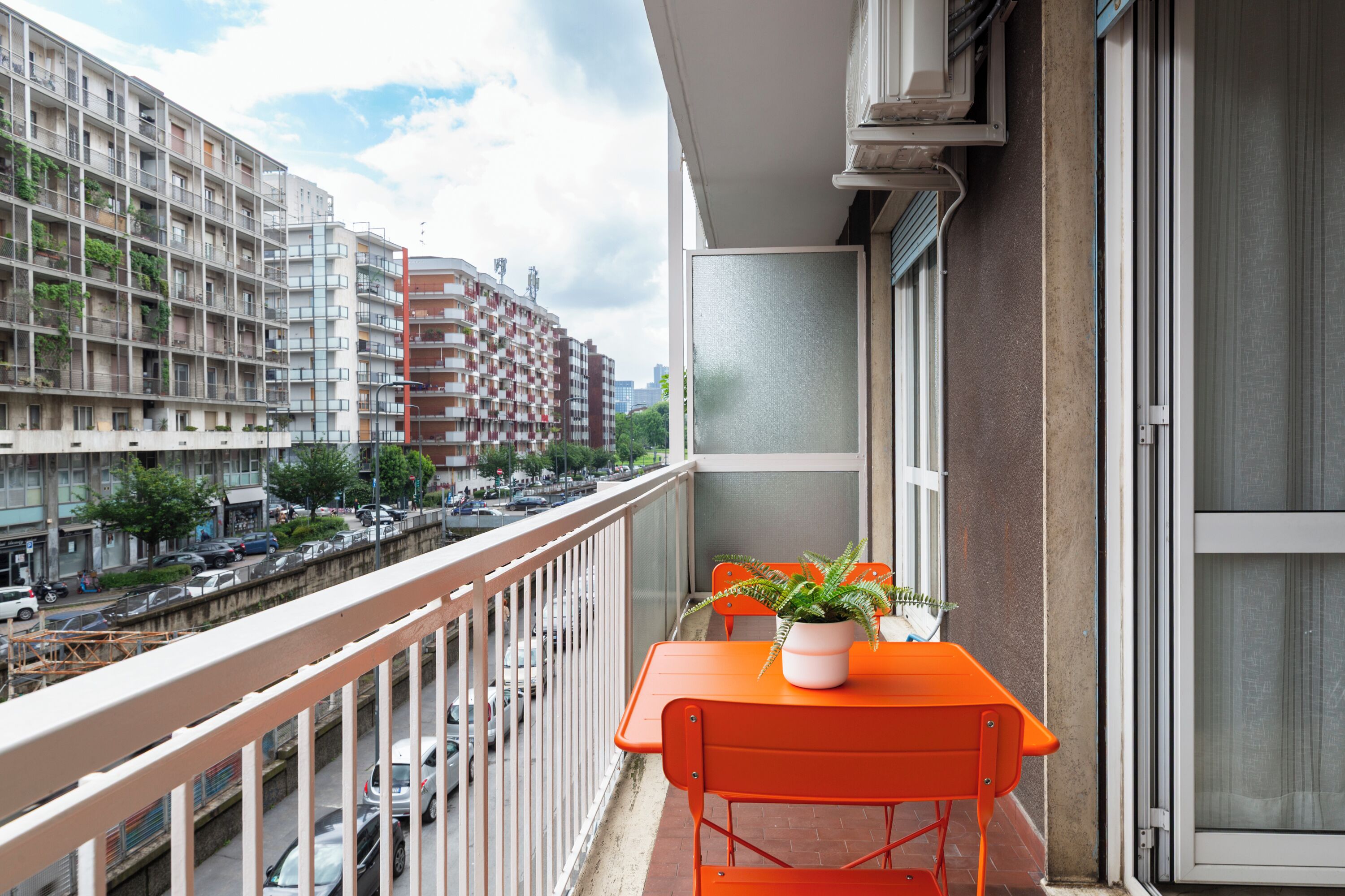 Un balcón con una mesa y sillas, una planta en maceta, y vista de una calle concurrida y edificios.