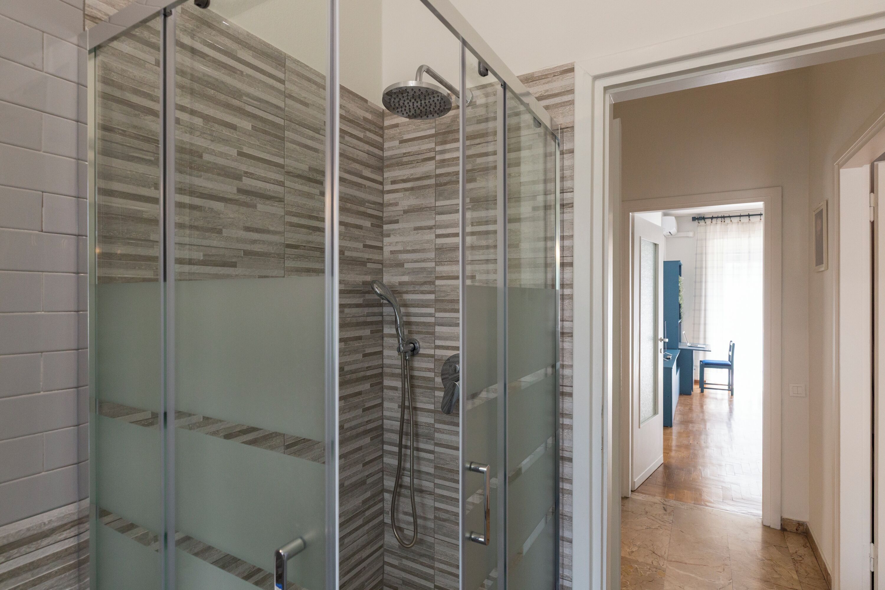 Un baño moderno con ducha envuelta en vidrio, paredes con azulejos y piso de madera que conduce a un comedor.