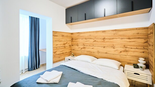A modern bedroom with a wooden headboard, a bed with white linens, a nightstand with a lamp, and a window with curtains.