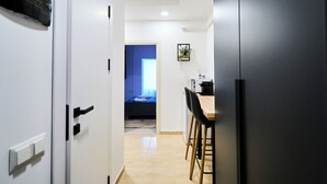 A hallway leading to a bedroom with a blue door, a small kitchenette with a bar stool, and a bed with a blue headboard.