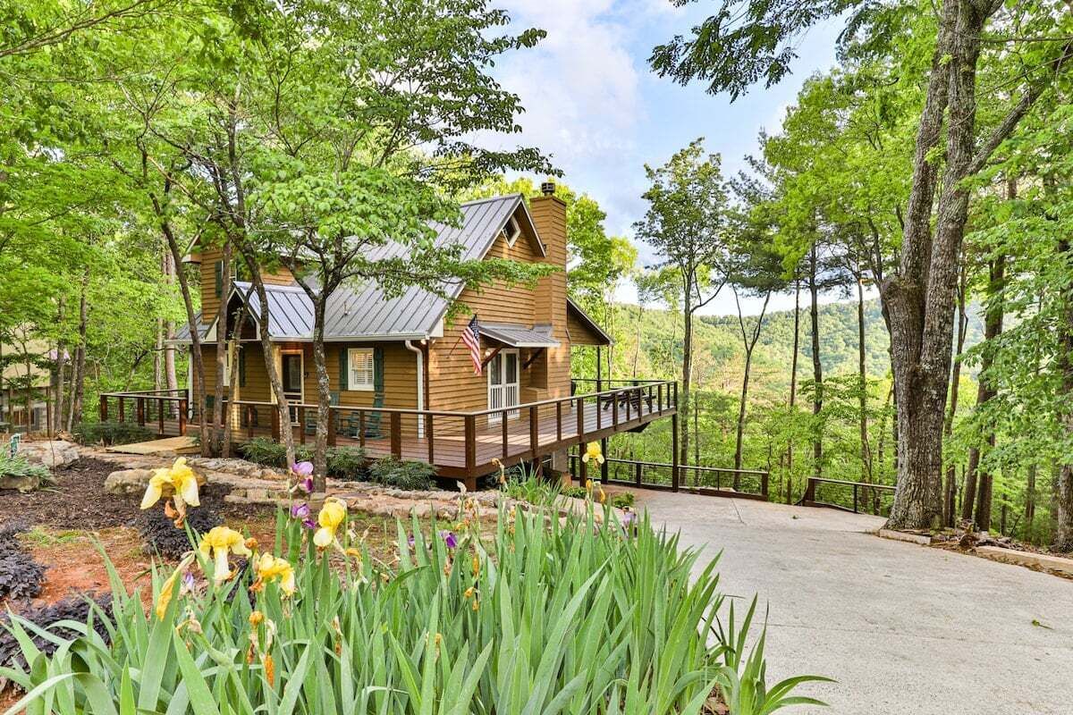 A wooden cabin with a metal roof, surrounded by lush greenery and a paved driveway.