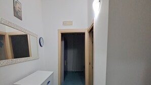 A narrow hallway with a white desk and a mirror on the left, a clock above it, and a door at the end.