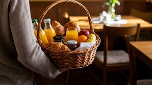 Ein Korbkorb mit Frühstücksartikeln, darunter Croissants, Obst und Orangensaft, gehalten von einer Person in einem grauen Pullover.