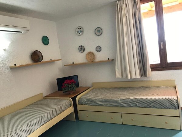 A small, cozy room with two single beds, a wooden nightstand with a TV and a vase of flowers, and a window with curtains. - Costa Smeralda Apartments (Cugnana Verde)