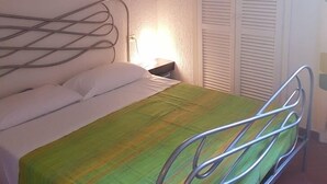 Ein Einzelbett mit Metallgestell, eine grün-gelb gestreifte Tagesdecke, weiße Kissen und ein Nachttisch mit Lampe.