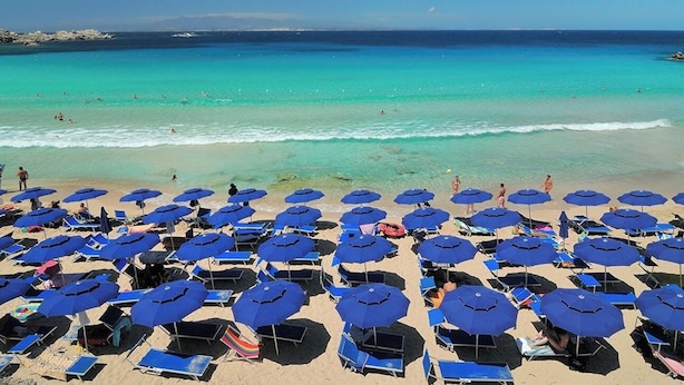 Ein Strand mit zahlreichen blauen Sonnenschirmen und Liegestühlen, einem klaren blauen Meer und ein paar Personen im Wasser.