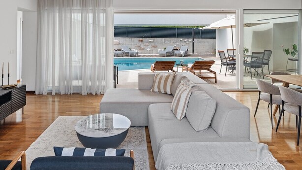 Ein modernes Wohnzimmer mit einem großen, grauen Ecksofa, einem Couchtisch und Blick auf einen Pool sowie eine Sitzgelegenheit im Freien.