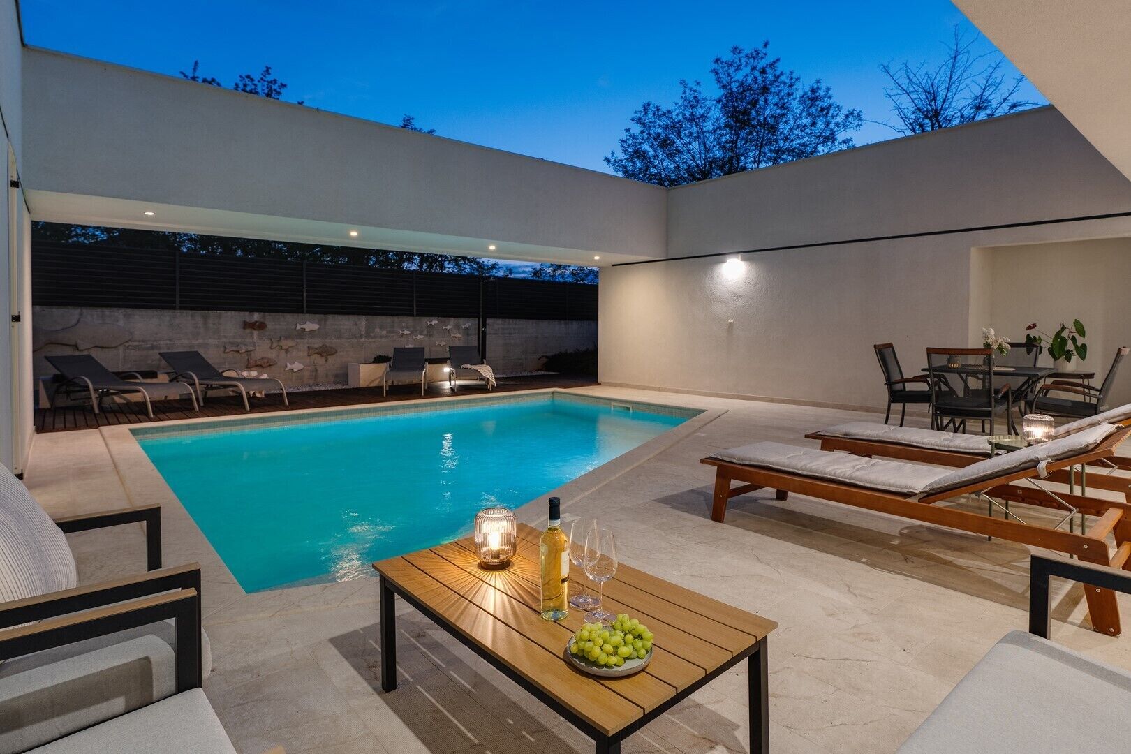 Zona de piscina exterior moderna amb gandules, taula de fusta amb ampolla i vasos, i taula de menjador amb cadires.