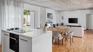 Eine moderne Küche mit Kochinsel, ein Essbereich mit Holztisch und -stühlen, ein Wohnzimmer mit Sofa und Fernseher sowie ein großes Fenster mit Vorhängen.