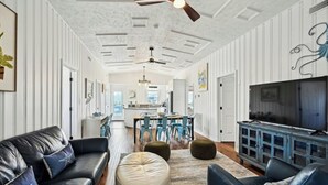 A spacious living room with a black leather sofa, a cream-colored ottoman, and a blue cabinet with a television. - Second Star- 7BR Channel House-Pool, Putting Green (North Myrtle Beach)