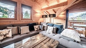 A cozy log cabin interior with a sectional sofa, a wooden coffee table, and large windows offering scenic views. - Solstice - Spacious Alpine Apartment Les Gets (Les Gets)