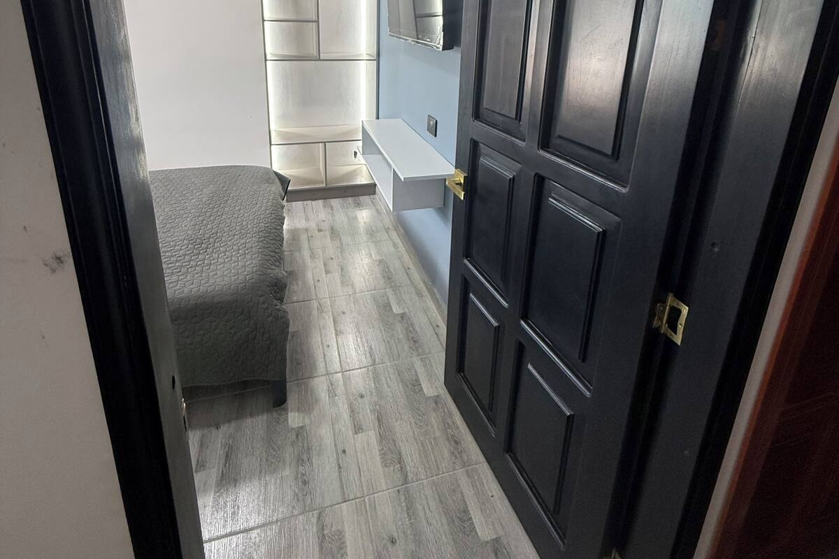 Une pièce avec une porte en bois foncé, une table de chevet blanche, un banc recouvert d’un tissu gris et un téléviseur fixé au mur.