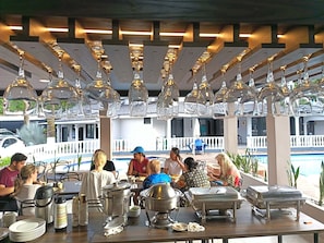 Un área de comedor al aire libre techada con mesas, sillas e lámparas de vidrio colgantes.