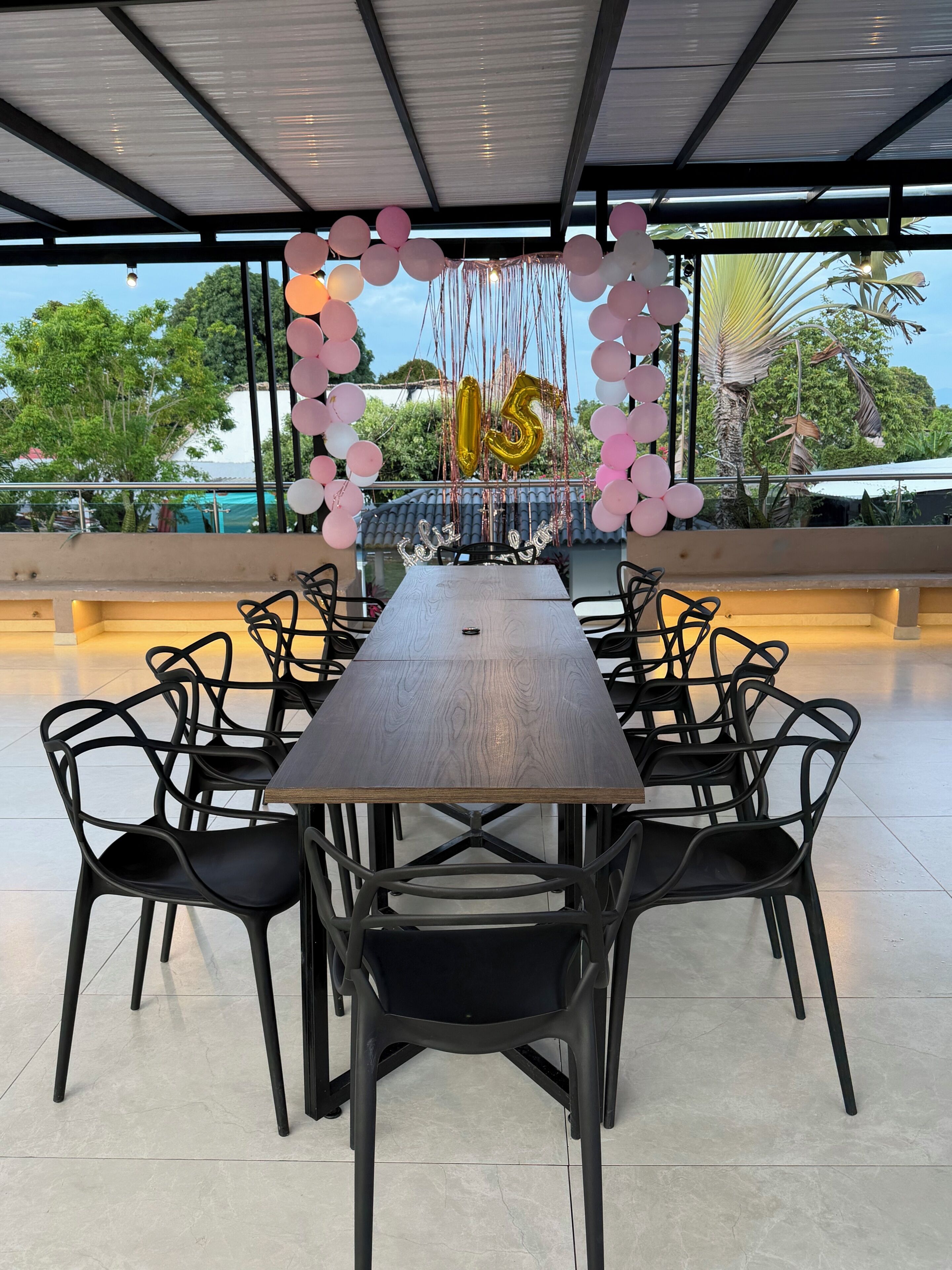 A long dining table with black chairs, surrounded by a decorative arch of pink and white balloons, and a golden '15' balloon.