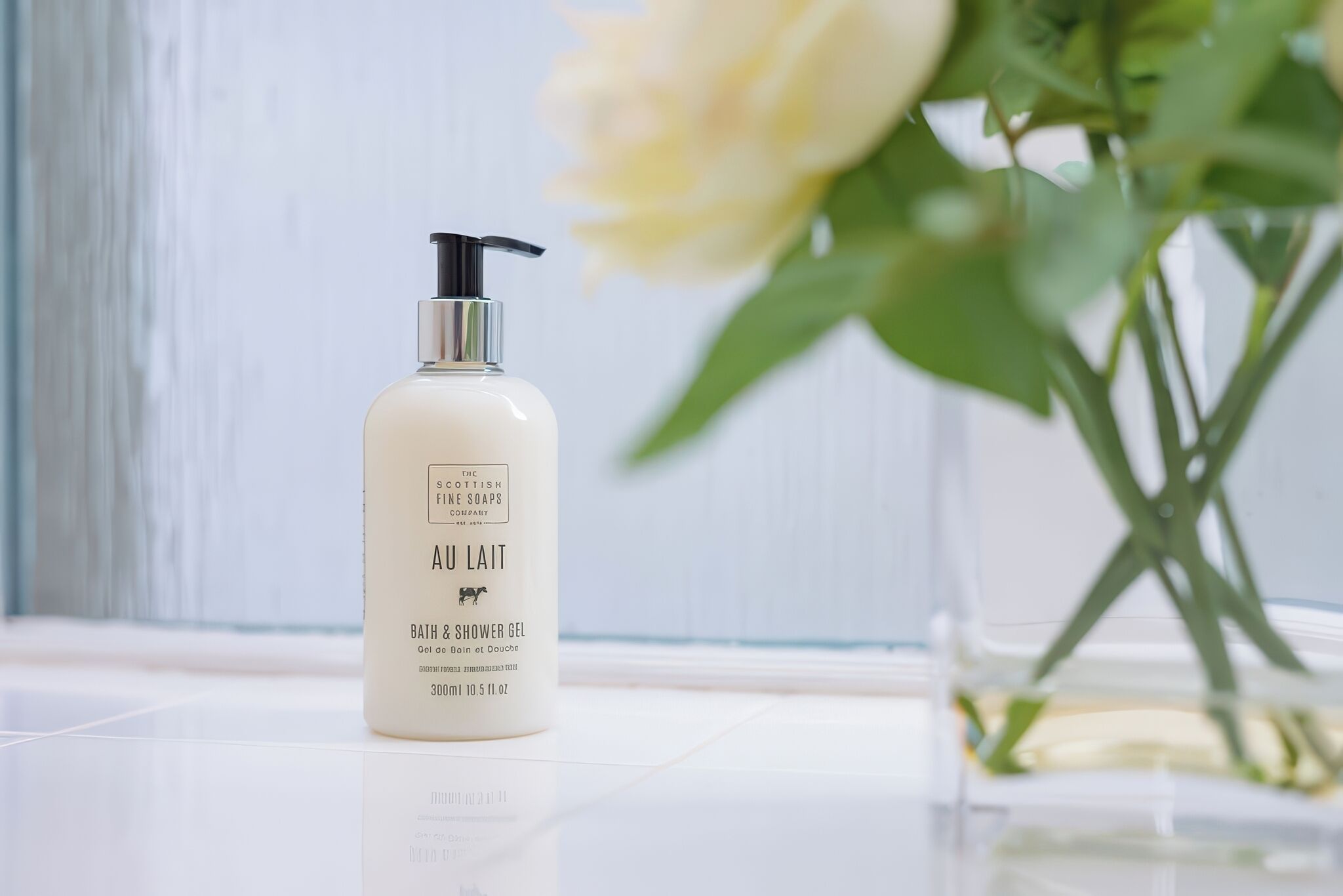 Eine Flasche Au Lait Bath & Shower Gel auf einer weißen Oberfläche, im Hintergrund eine weiße Wand und eine Blumenvase.