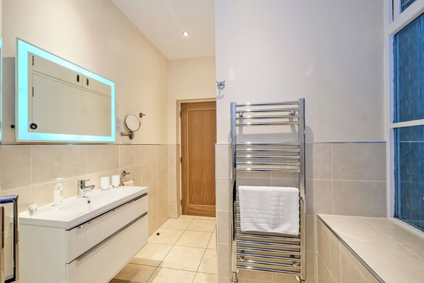 A modern bathroom with a large mirror, a sink, a towel rack, and a window. - Tullich Apartment (Ballater)