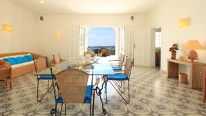 A dining area with a glass table and chairs, a sofa with blue cushions, and a view through open doors.