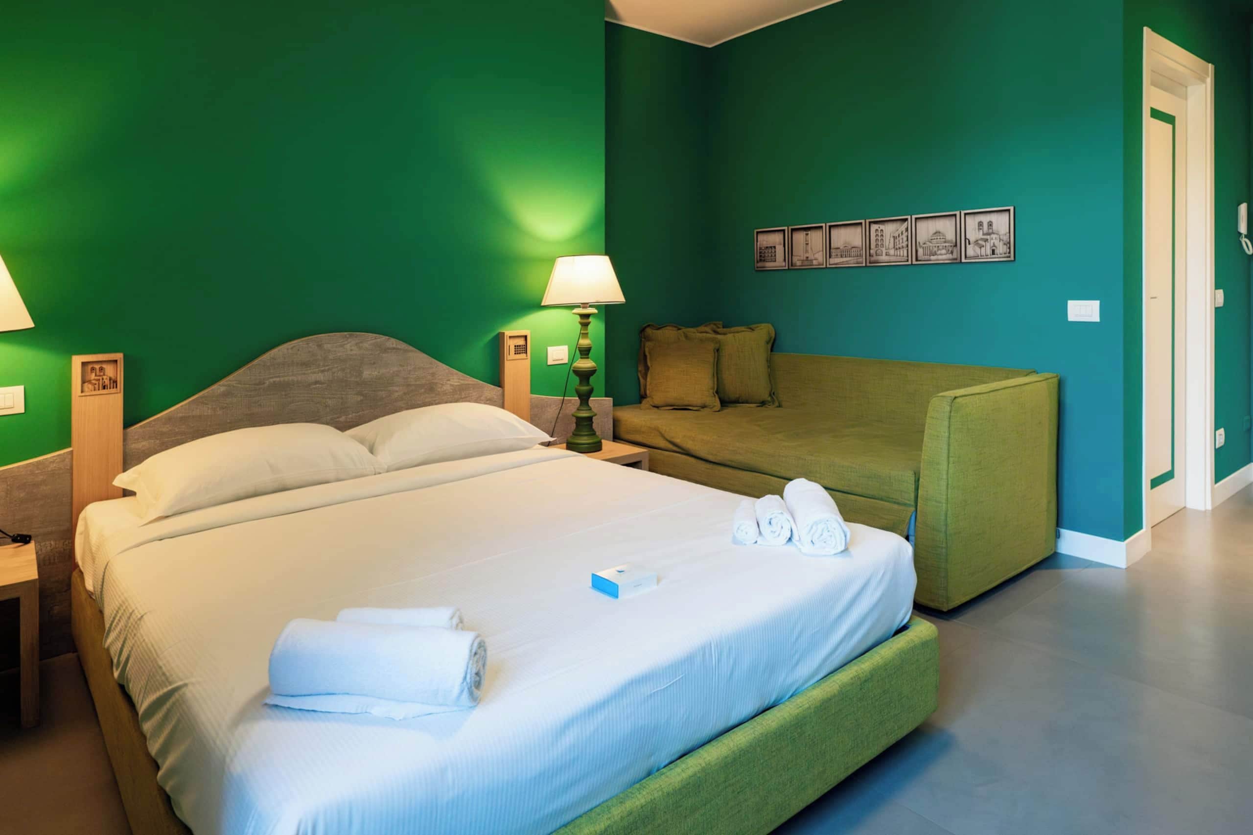 Una habitación de hotel con una cama, un sofá verde, una mesita de noche con una lámpara y una pared con cuadros enmarcados.