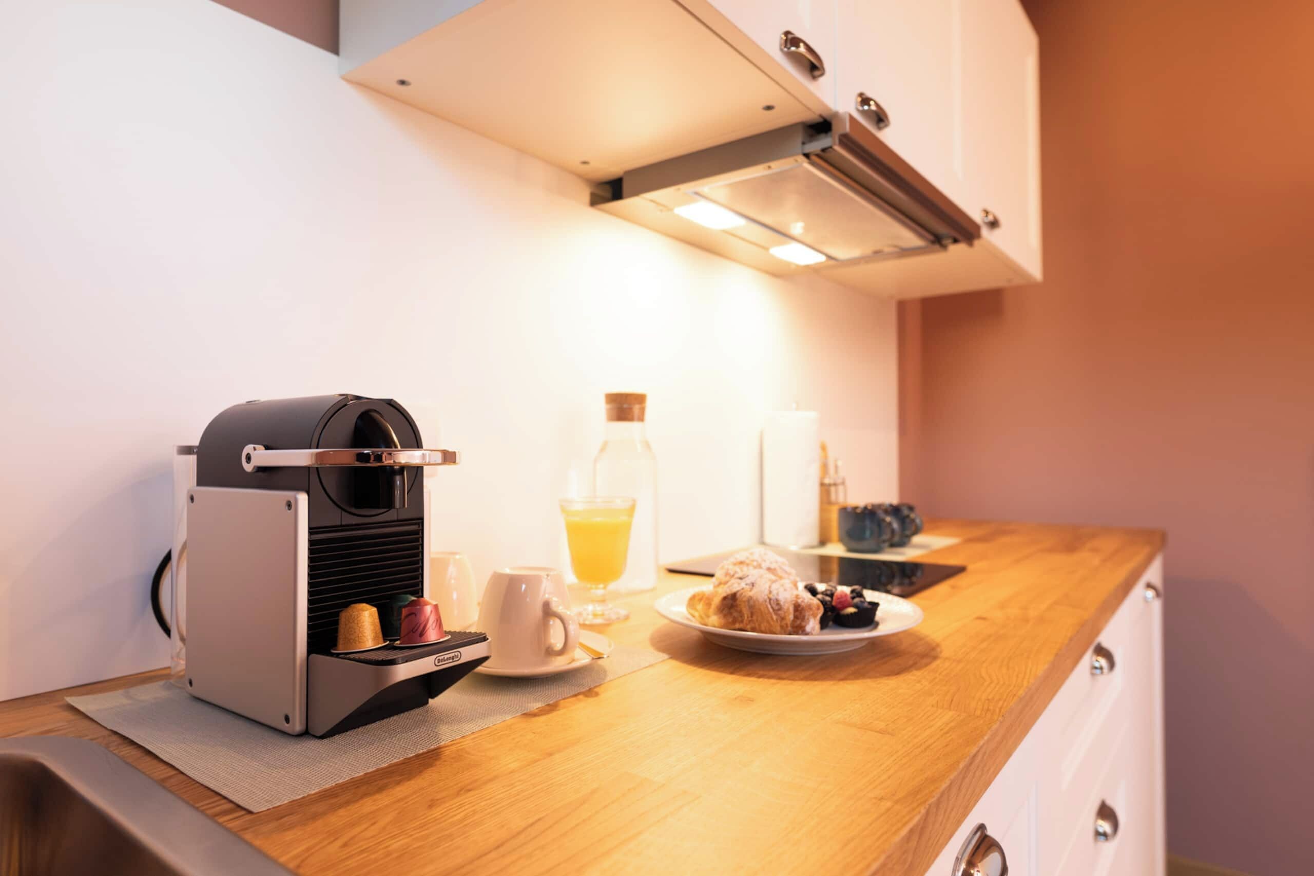 A kitchenette with a coffee machine, a glass of orange juice, a plate with pastries, and a coffee cup.