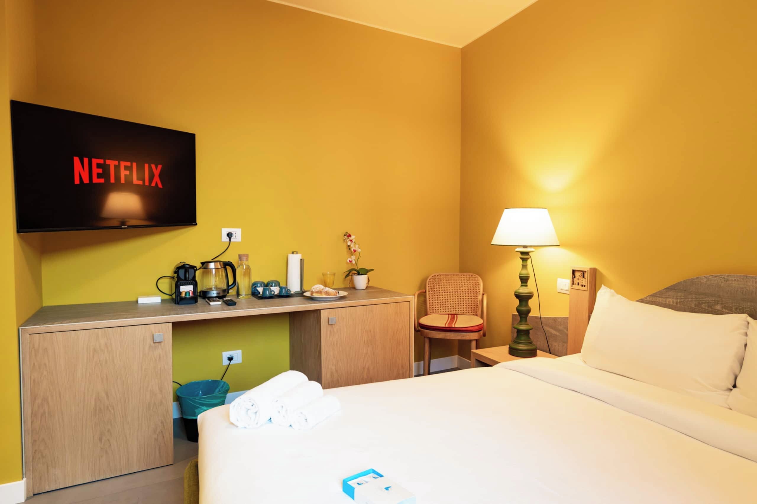 A hotel room with a bed, a desk with a coffee maker, a lamp, and a TV displaying the Netflix logo.