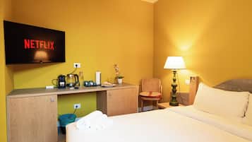 A hotel room with a bed, a desk with a coffee maker, a lamp, and a TV displaying the Netflix logo.