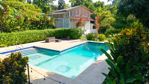 A swimming pool with a tiled deck, surrounded by lush greenery and a house with a satellite dish.