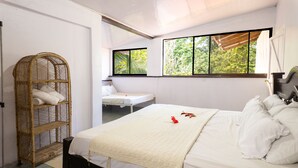 A room with a bed, a small table, a wicker chair, and a window with a view of greenery.