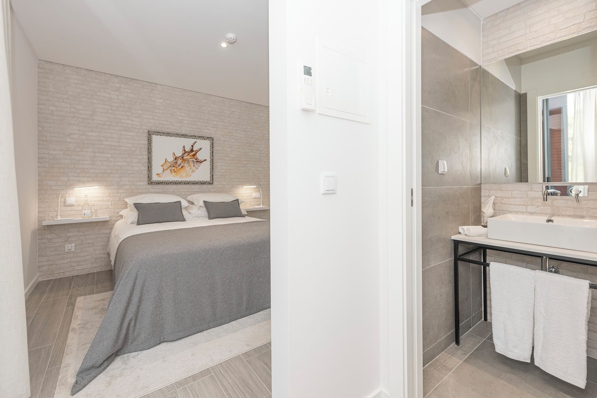 Un dormitorio moderno con cama, lámparas de noche, aire acondicionado montado en la pared y baño con lavabo y espejo.