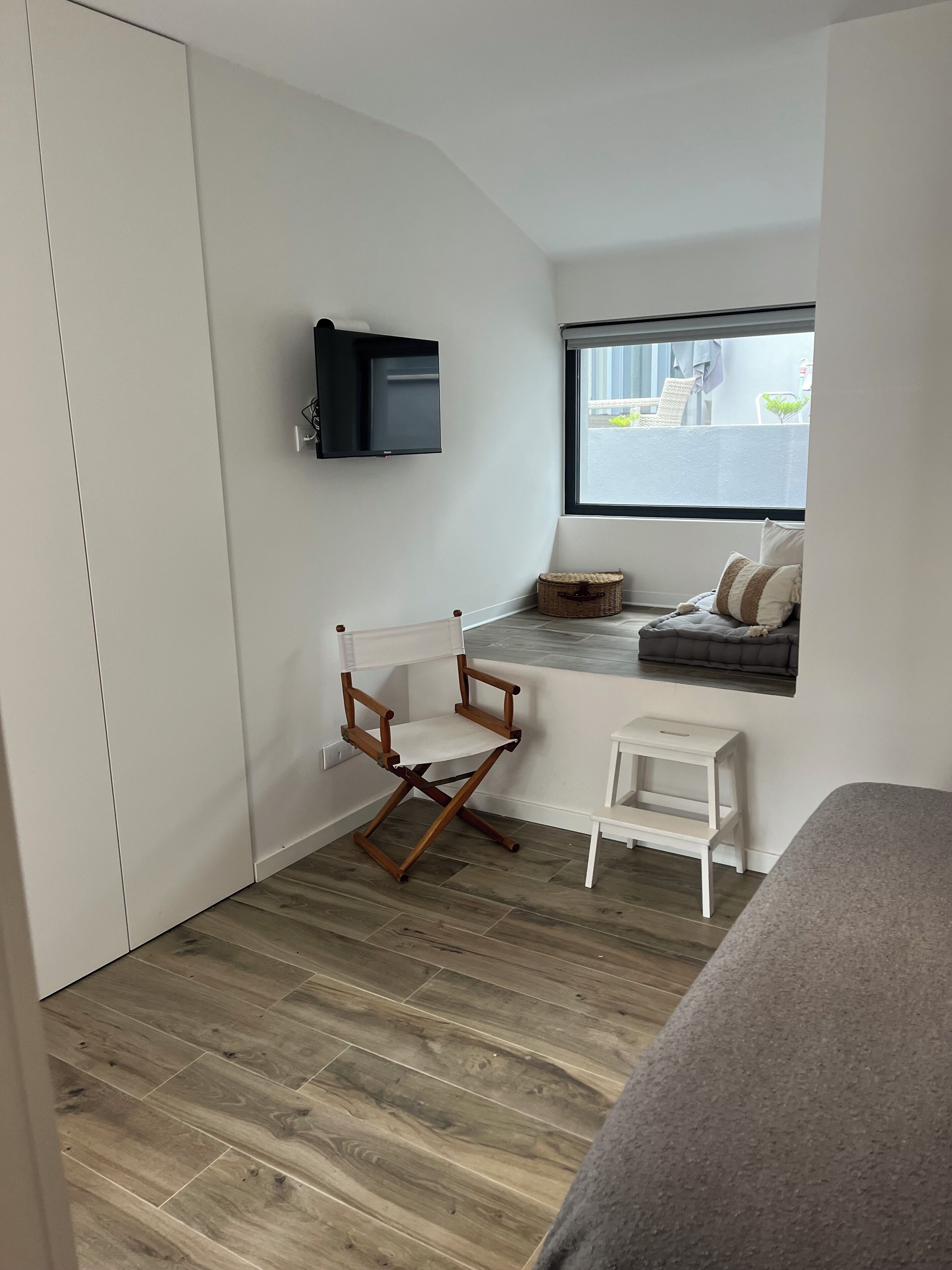 A compact room with a wooden floor, a small white desk, a wooden folding chair, a television mounted on the wall, a window with a view of the outside, and a bed with a grey cover.