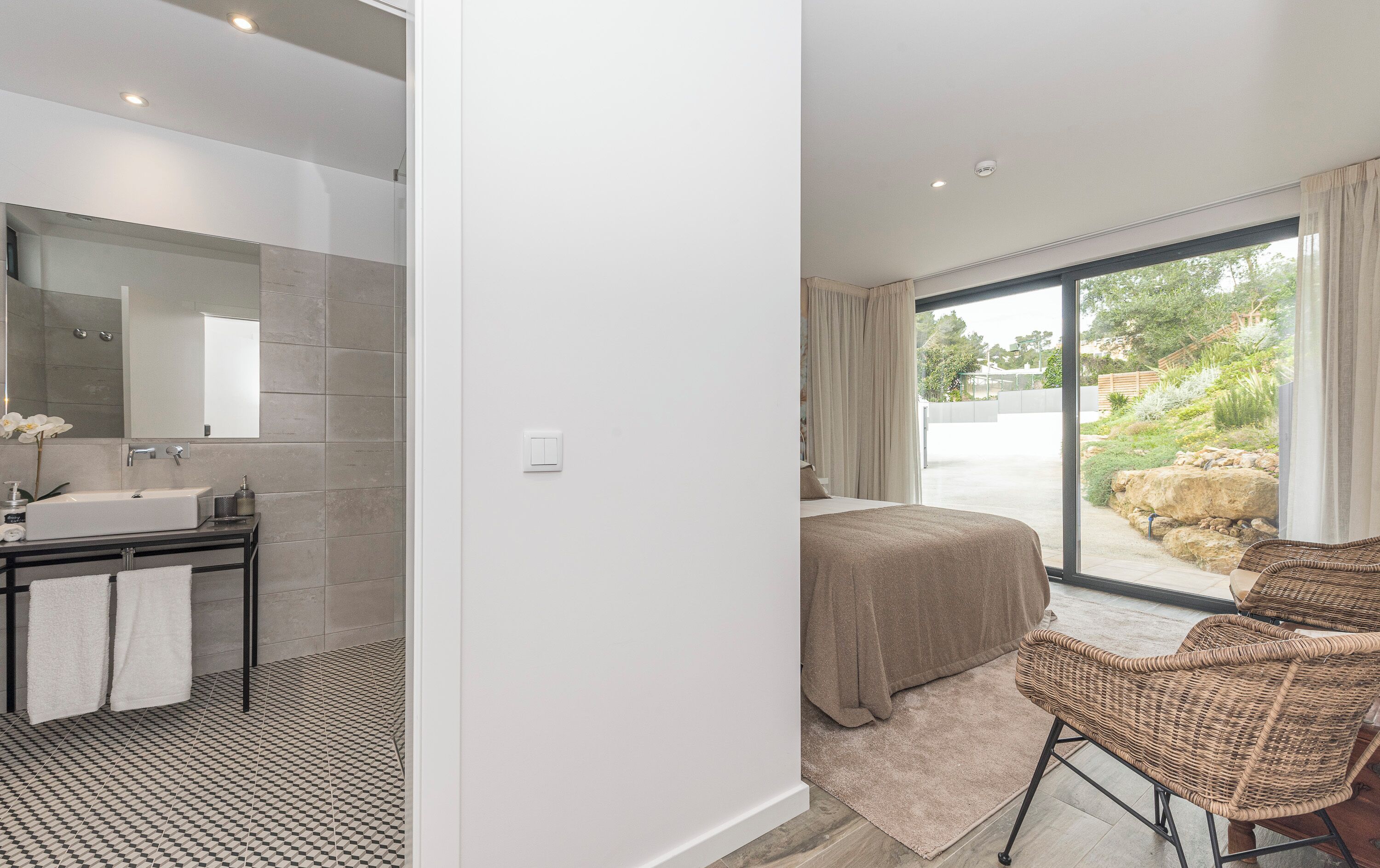 Um banheiro moderno com um espelho grande, pia e chuveiro. Há uma cama com vista para o exterior através de uma porta de vidro.