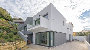 Una casa moderna de dos pisos con balcón, amplias puertas correderas de vidrio y un muro de contención de piedra.