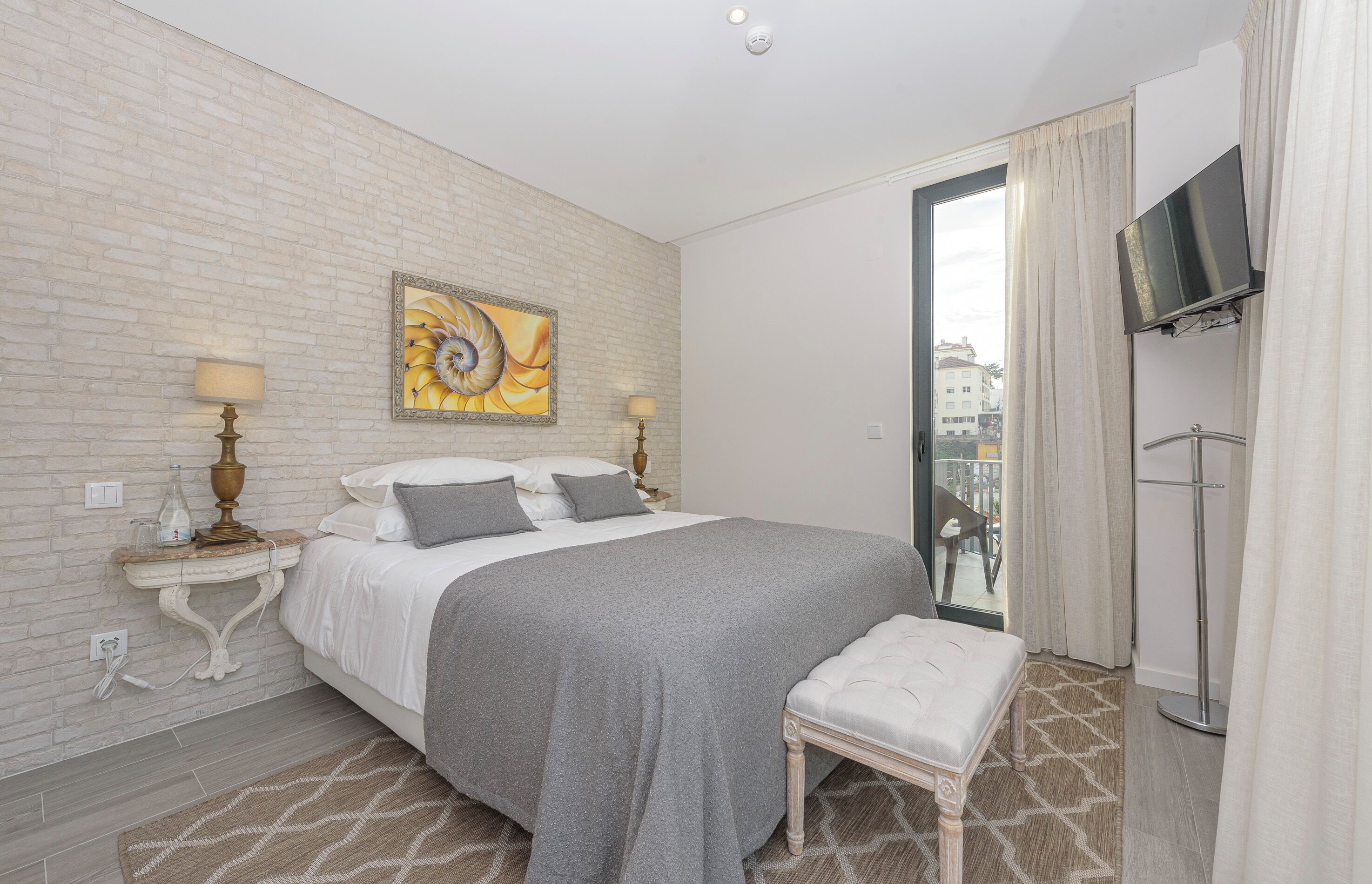 Habitación de hotel con una cama grande, un televisor de pantalla plana montado en la pared, una mesita de noche con una lámpara y vistas a la ciudad a través de un ventanal amplio.