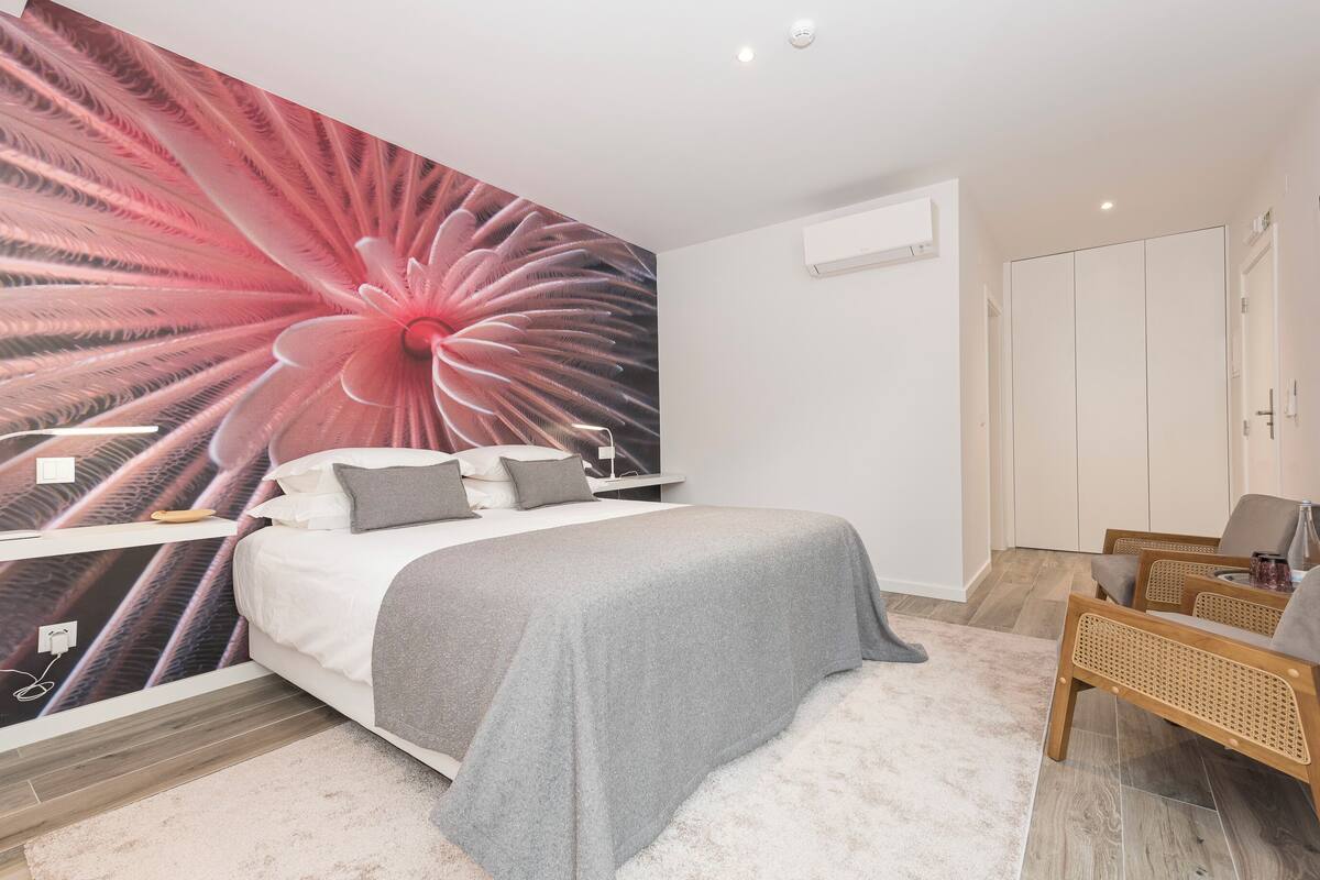 Un dormitorio con un mural grande en la pared, una cama con ropa de cama gris, una mesita de noche con una lámpara, una silla y un televisor montado en la pared.
