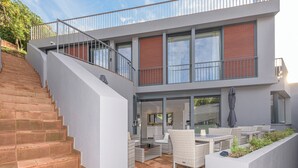 Una casa moderna con una escalera que lleva a un balcón, un patio con muebles de exterior y ventanas amplias.