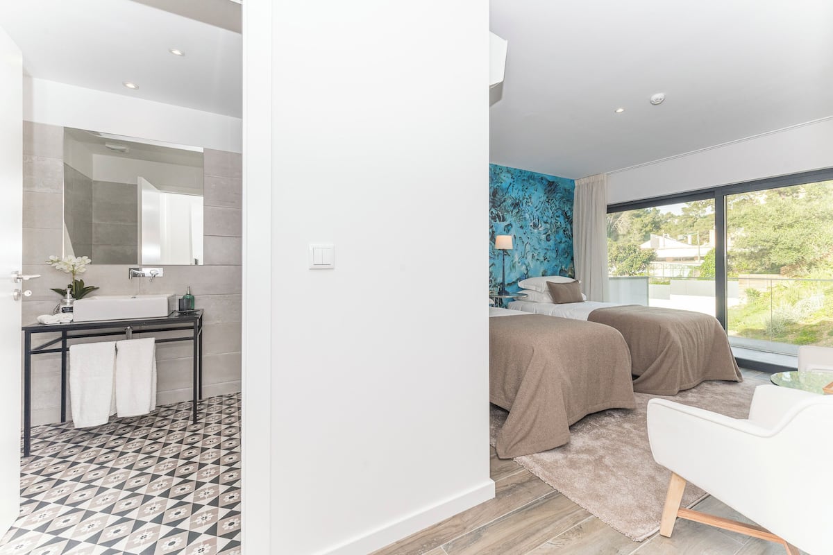 Un baño moderno con un espejo grande, lavamanos y un perchero para toallas. El baño tiene un piso estampado y una ventana amplia con vistas a la vegetación.