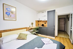 A bedroom with a bed, pillows, a green chair, a small table with a plant, and a kitchen area with cabinets and a coffee machine. - Studio Near Airport • Fast Check-In (Chișinău)