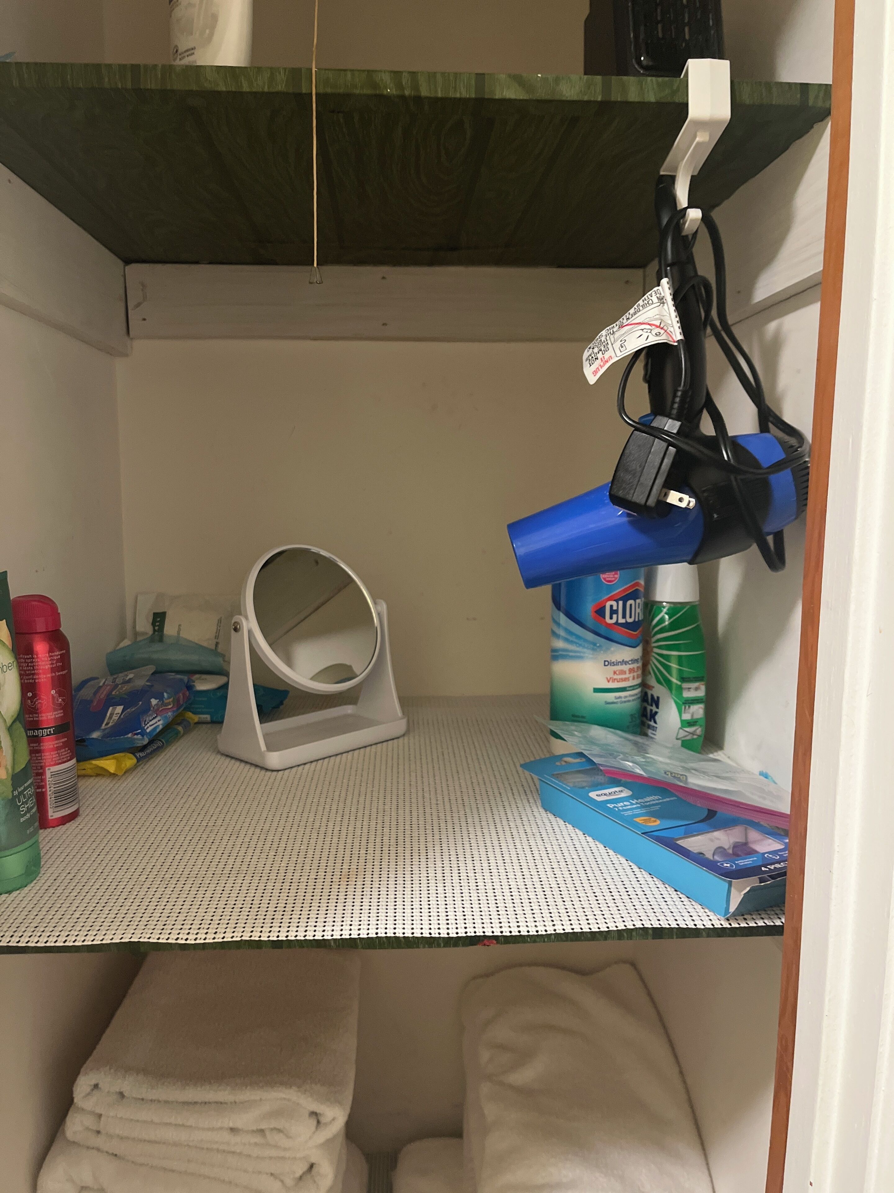Un espacio de almacenamiento pequeño con un estante blanco que contiene un pulverizador azul, una lámpara blanca y diversos productos de limpieza.
