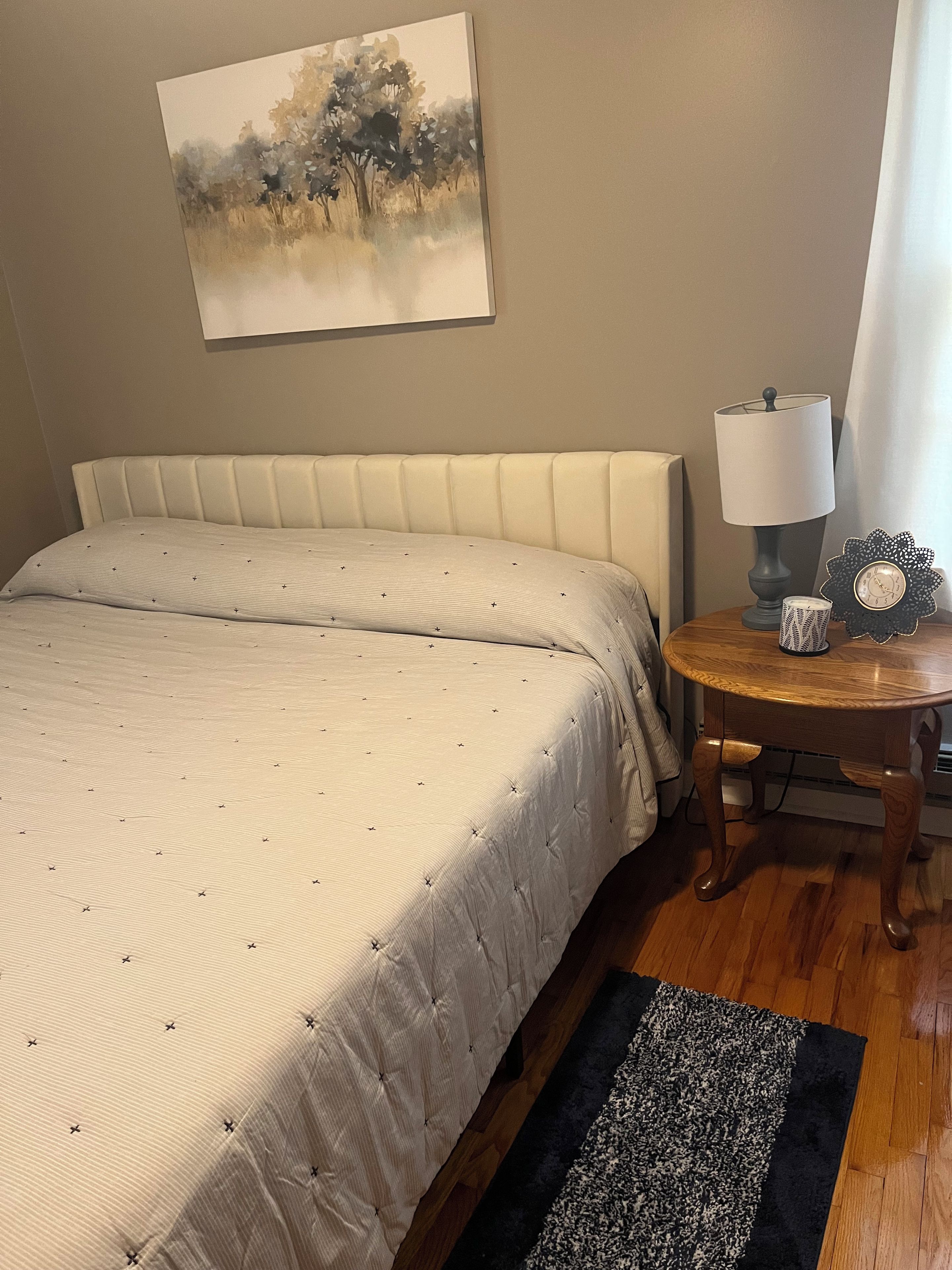 A bedroom with a bed, a bedside table with a lamp, a clock, and a painting on the wall.