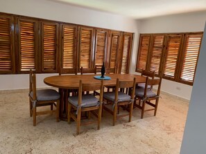 Un comedor con una mesa redonda de madera rodeada por seis sillas, cada una con un cojín. La habitación tiene ventanas grandes con contraventanas de madera.