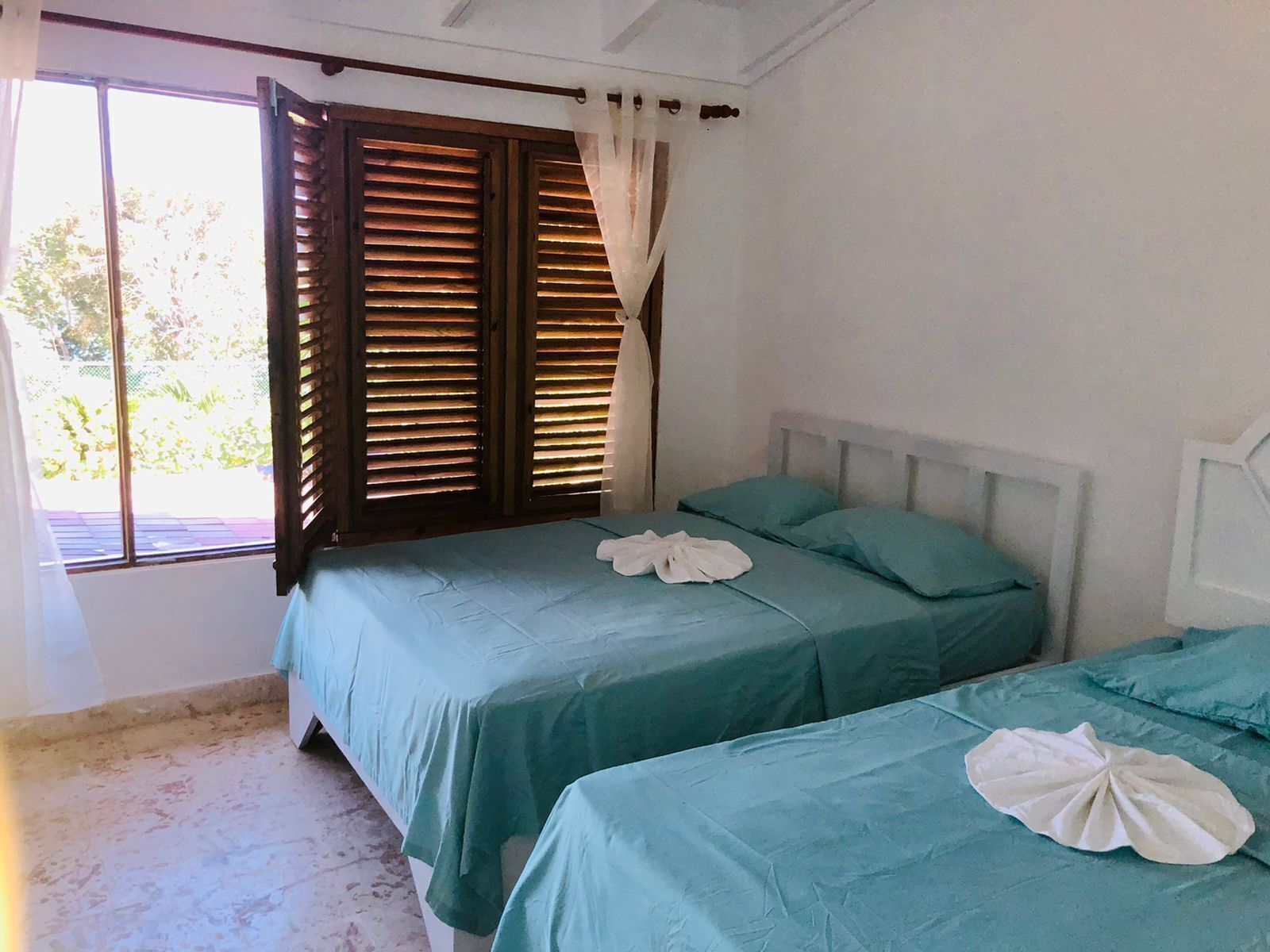 A room with two beds, each with a teal bedspread and white pillows. There is a window with wooden shutters and a curtain.