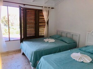 Una habitación con dos camas, cada una con una colcha color turquesa y almohadas blancas. Se observa una ventana con contraventanas de madera y una cortina.