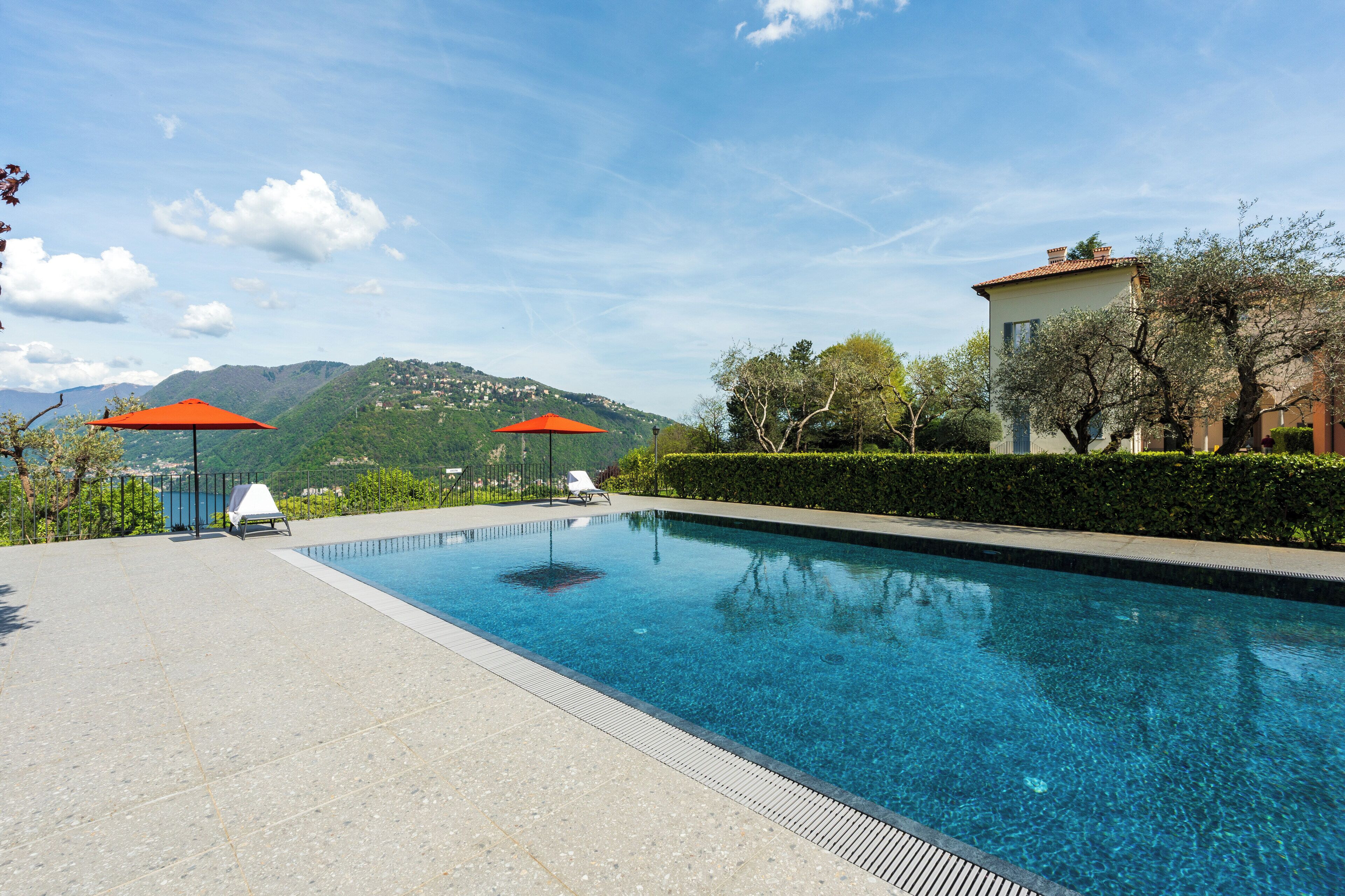 A swimming pool with two sun loungers and umbrellas, surrounded by a landscaped area with a view of mountains.