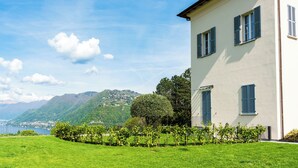 A building with a white facade and blue shutters, surrounded by a well-maintained lawn and a view of a lake and mountains. - Villa Maderni Breathtaking (Como)