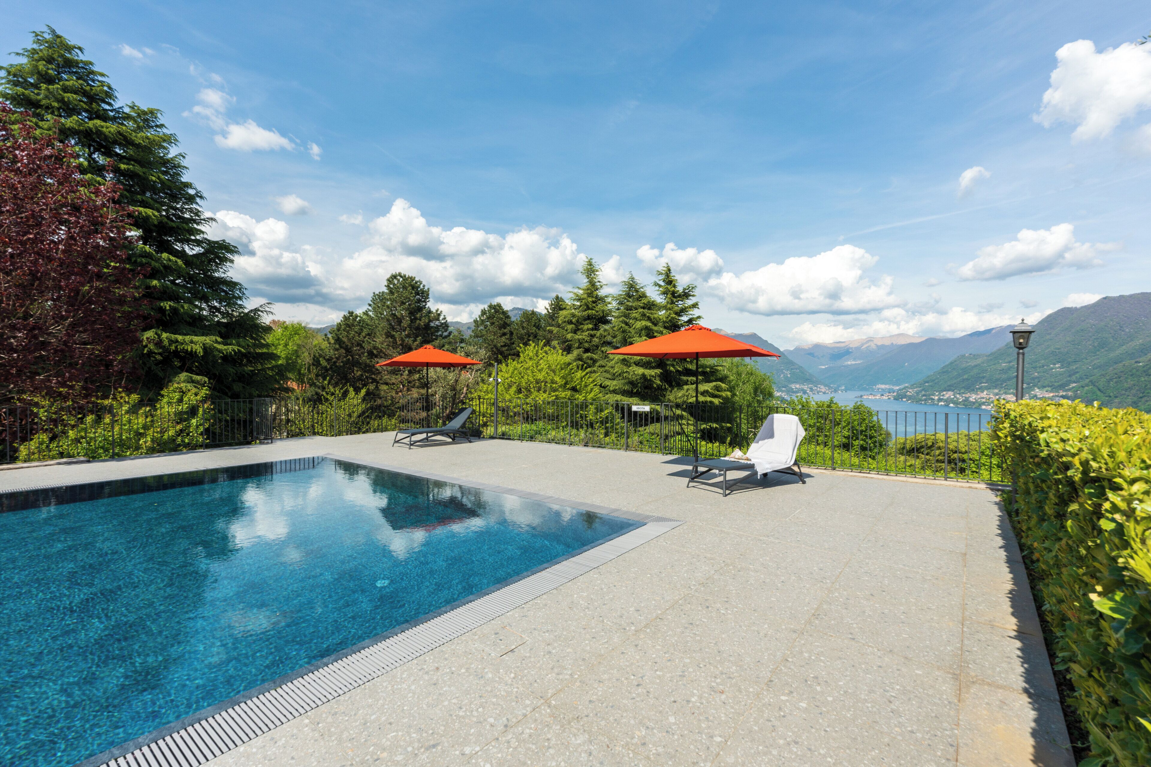 Ein Schwimmbecken mit zwei roten Sonnenschirmen, einer weißen Liege und Blick auf Berge und einen See.