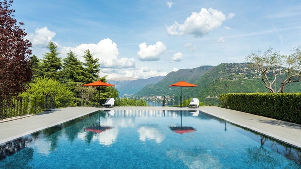 A clear swimming pool with two umbrellas, surrounded by greenery and mountains in the background. - Villa Maderni Breathtaking (Como)