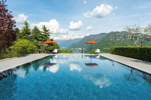 A clear swimming pool with two umbrellas, surrounded by greenery and mountains in the background.