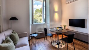 A modern living room with a sofa, a dining table with chairs, a TV mounted on the wall, and a window with an open view. - Villa Maderni Breathtaking (Como)