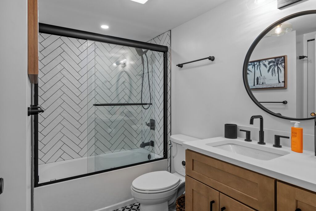 Une salle de bain moderne avec une douche carrelée en motif chevron, des toilettes blanches, un meuble-lavabo en bois et un grand miroir.